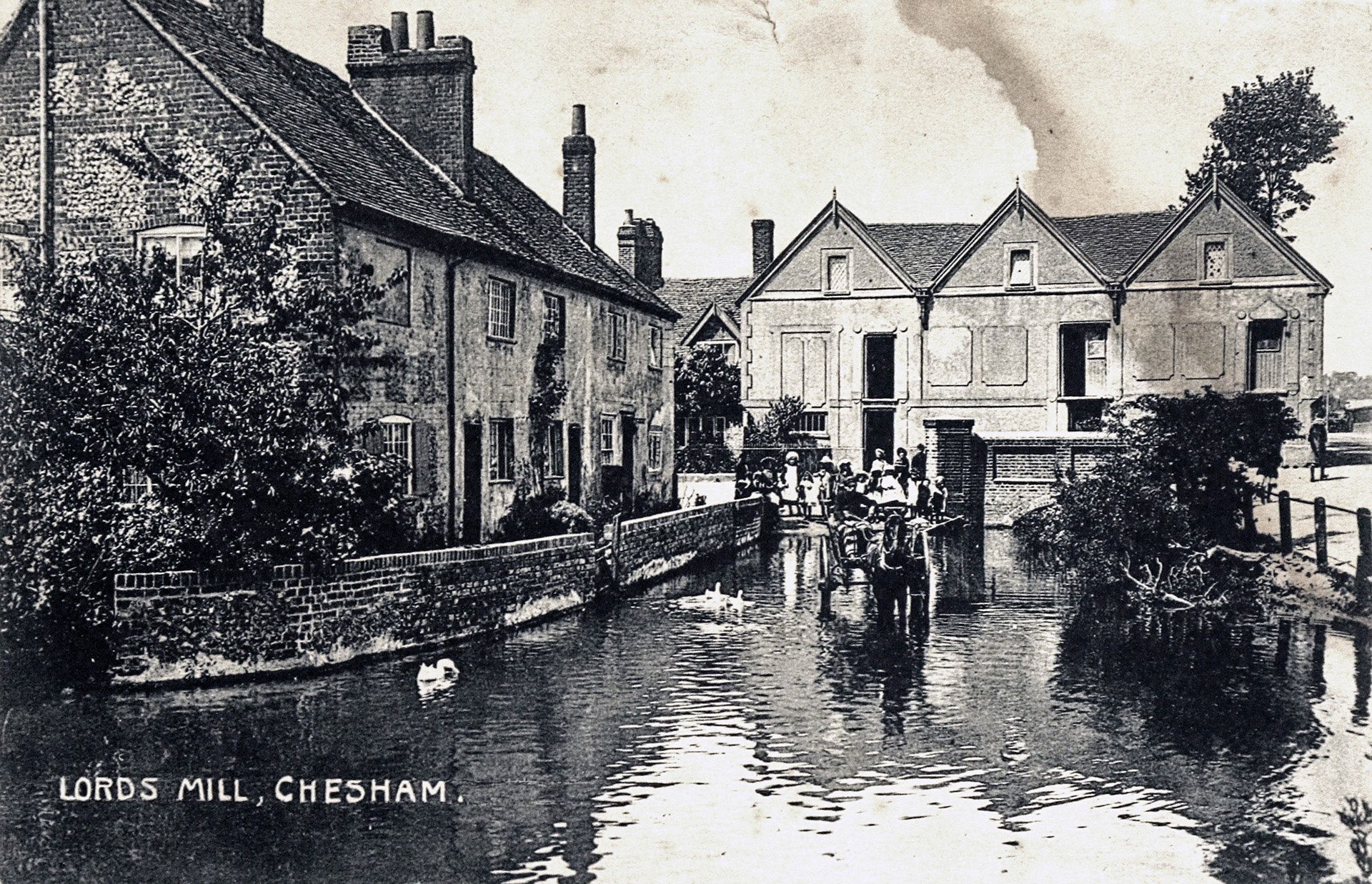 A black and white postcard depicting Lords Mill. Houses surround the water with a horse and cart carrying people travel through.