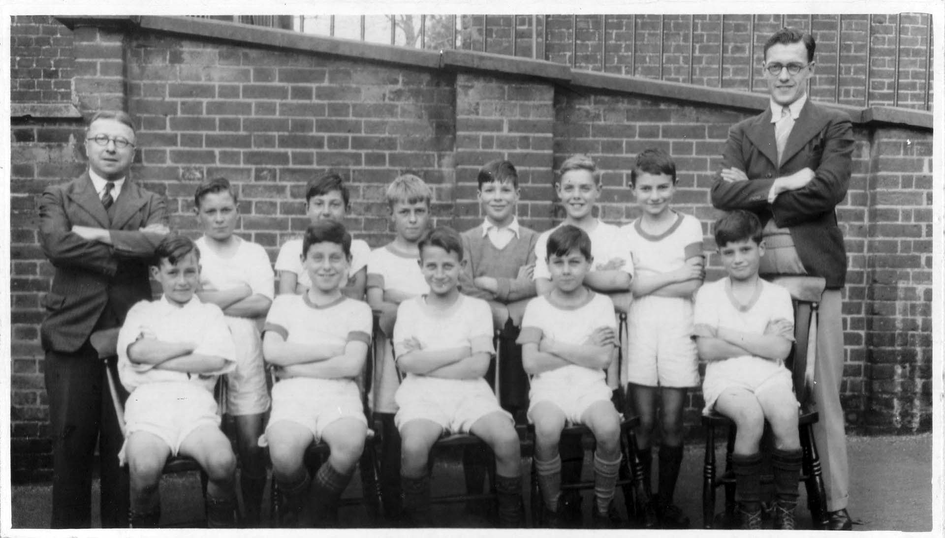 Black and white photo of 11 children seated in white tops and shorts with 2 teachers standing either side