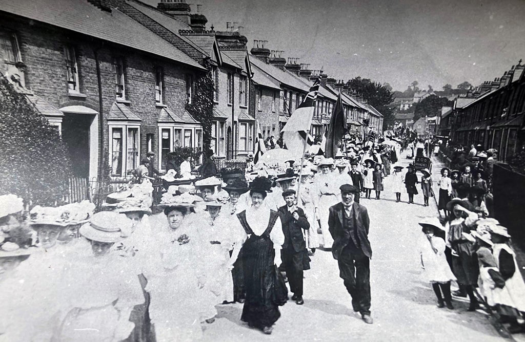 Men and women wearing hats walk down the street followed by children