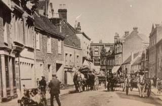 High Street, Chesham. There are various buildings, some of which are shops. People are on the street and there are horses pulling carts.