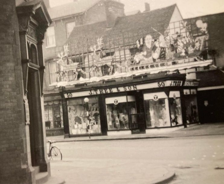 Black and white photo of a shop which has a sign that says 'J Tree & Son'. The shop has the number 50 on it and its three window displays show various clothing items. There are Christmas decorations above the shop frontage.