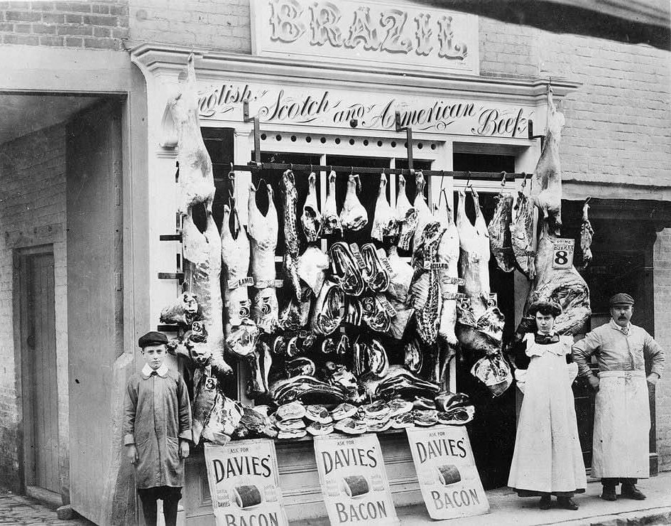 A photo of Brazil butchers shop showing various meats hanging from hooks on a rail attached to the outside of the shop front. A man, woman and child stand outside next to three signs that state 'Davie's Bacon'