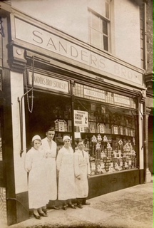 Two women, one man and a child are wearing aprons and standing outside a shop that has a sign above the shop saying 'Sanders Bros Ltd'