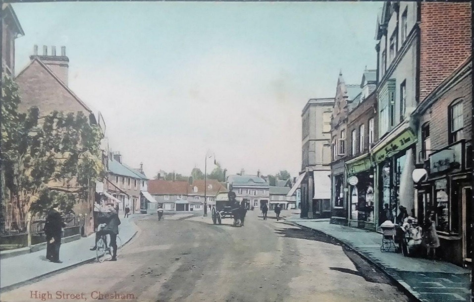 Postcard–titled 'High Street, Chesham'–showing high street buildings with a large unpaved street in the middle used by horse and cart. Pavements either side have people walking, pushing a pram or stopping to chat with their bicycle.