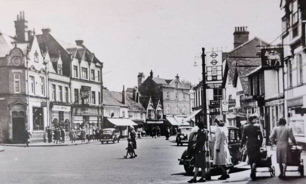 Photo of Chesham High Street showing the bakers. There is a bus stop in the forefront with people waiting. Cars are nearby and people are walking on the pavements next to the shops