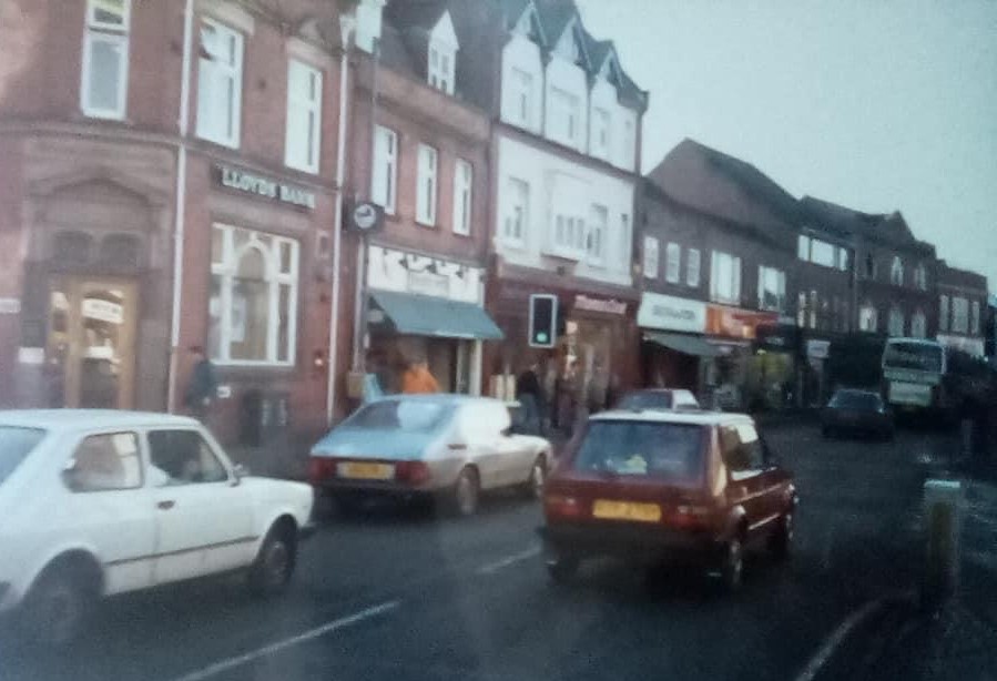 Colour photo of the High Street. There are cars travelling down the road next to shops and Lloyds Bank on the corner