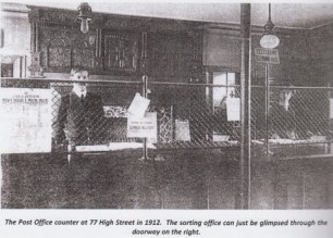 The interior of the post office showing two staff, a man and a woman, stood behind a wire barrier. The text on the image says: "The Post Office counter at 77 High Street in 1912. The sorting office can just be glimpsed through the doorway on the right."