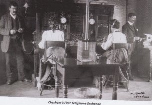 Chesham's first telephone exchange. The photo shows two women working at the exchange. They are both seated and wearing headsets. Two men stand either side.