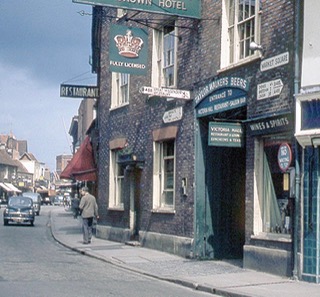 Colour photo of the front of The Crown, a brown bricked building with green entrance and sign with a crown on it which hangs from another sign. A man walks past and cars can be seen on the road