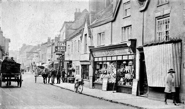 Black and white photo of the High Street showing shop fronts, including Easts Stationers. There are horses with carts travelling up the street and people walking on the pavement.