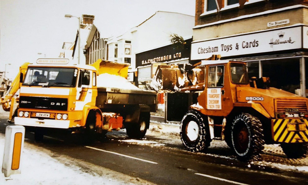 Two large vehicles are being used to clear snow from Chesham Broadway in 1982. A sign for Chesham Toy and Cards shop can be seen behind them