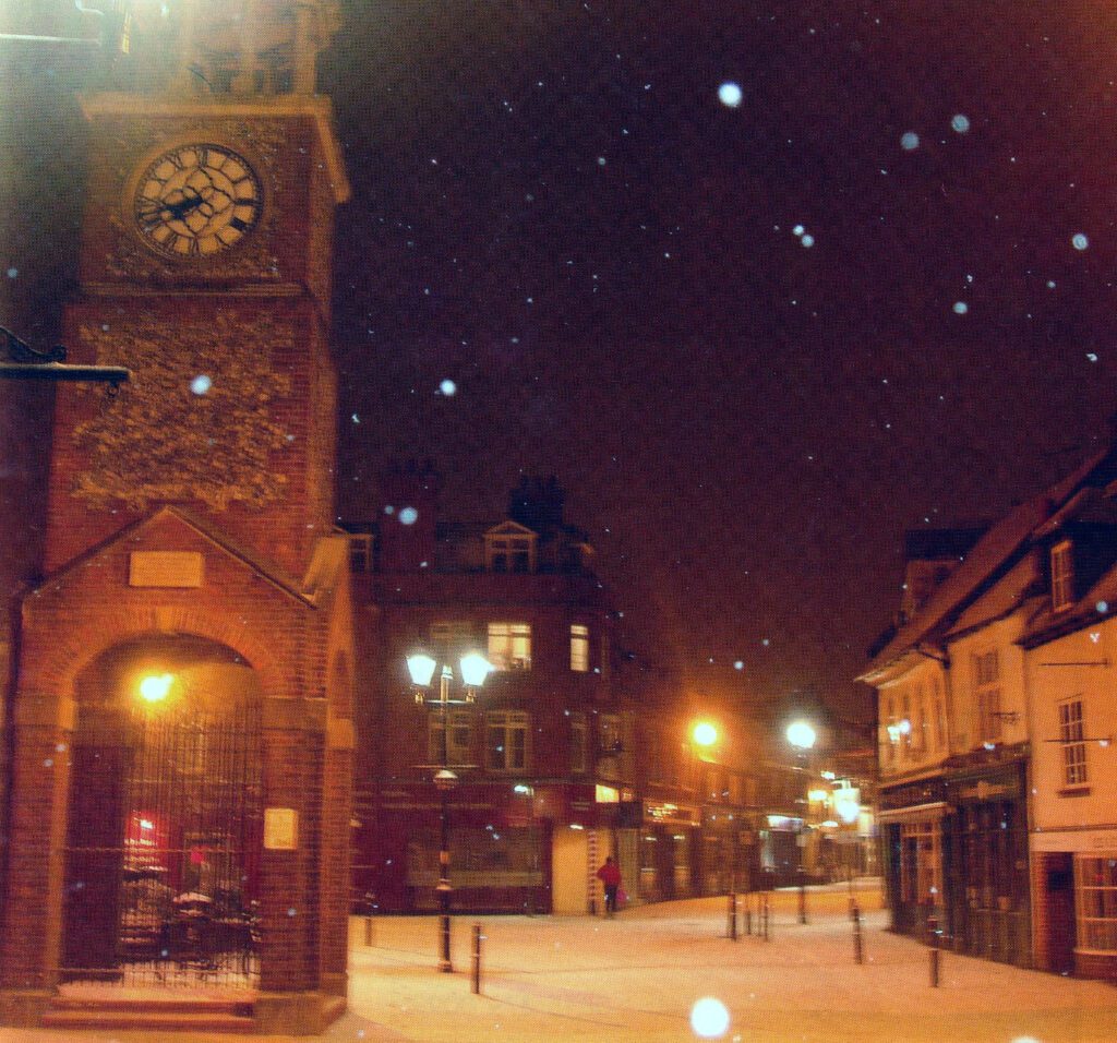 A photo of Market Square. It is snowing and snow lies on the ground. The clocktower is prominent on the left and other premises on the square are visible