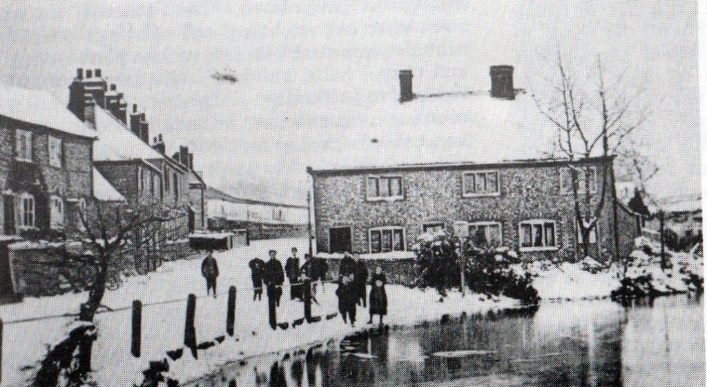A black and white photo of Waterside in 1925. There is snow everywhere, including roof tops. People gather next to the river. There are houses behind them.