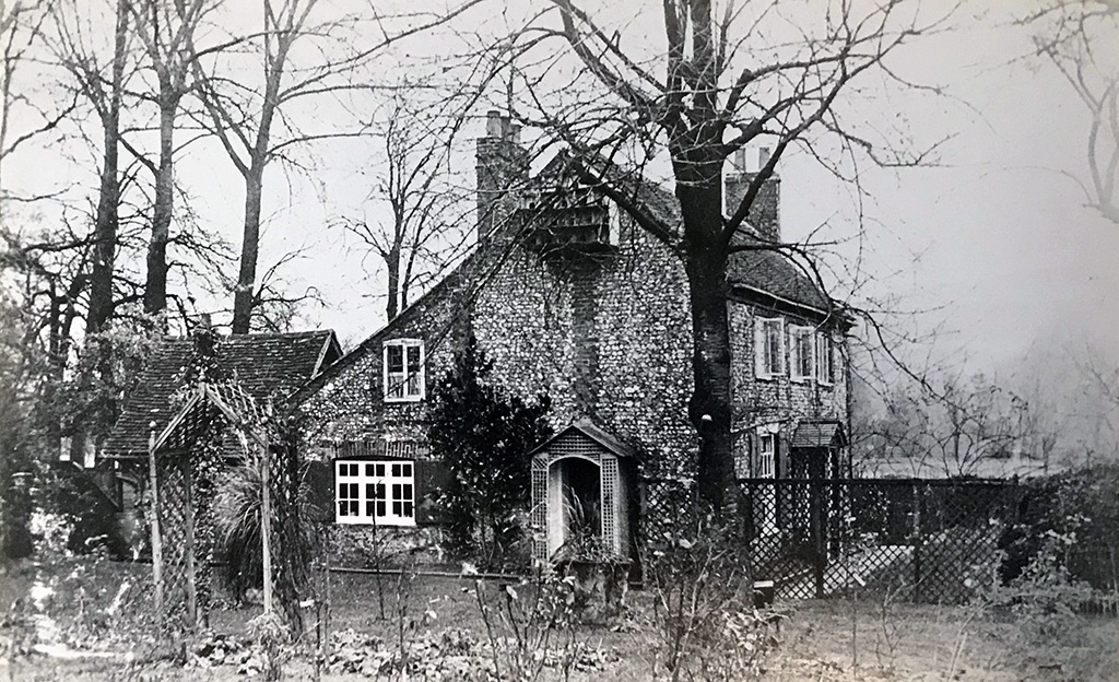 Black and white photo of The Cottage Amy Lane. There are trees surrounding the cottage and a wooden archway at the fence at the entrance of the garden