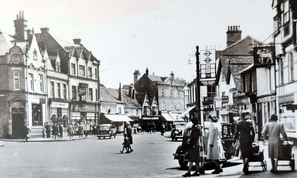 A black and white photo of Chesham High Street with some people waiting at a bus stop and others strolling down the footpaths past shops