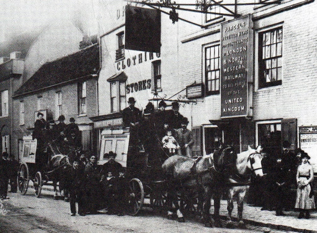 The horse bus to Watford - there are 2 coaches with horses positioned outside a store called Clothing Stores. Passengers are at on the coach with other people stood by looking on.