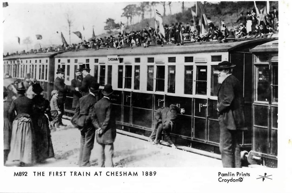 Steam train at Chesham Station. People stand on the platform observing. A man is seen adjusting the step onto the train. 