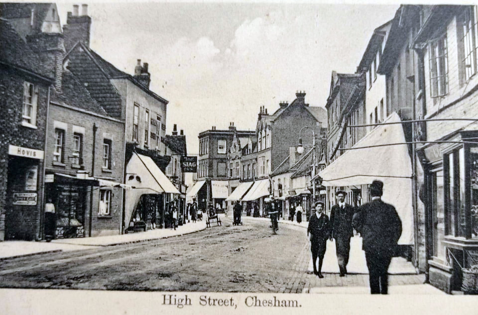 Black and white photograph of High Street, Chesham. Shops are on either side of the road with people walking up and down.