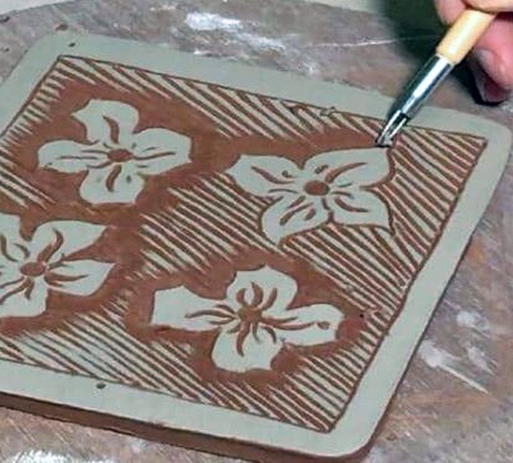 An artist uses a carving tool to create a floral design on a square piece of clay using the sgraffito technique. The clay is a light grey color, and the carved-out areas reveal a reddish-brown layer underneath. The design features four stylized flowers, each with four petals, set against a background of diagonal lines.