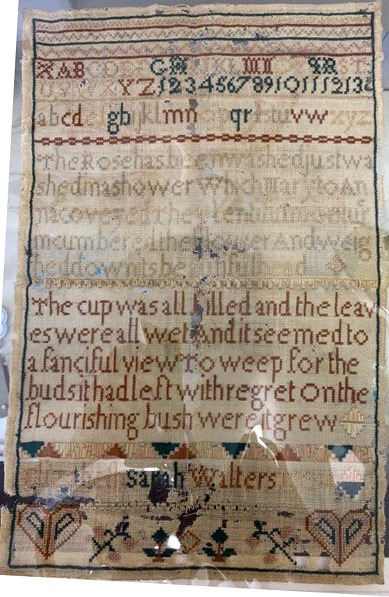 An antique sampler featuring various stitched letters, numbers, and a poem. The top section includes uppercase and lowercase letters along with numbers 1 to 13. Below the letters is a decorative horizontal border. The middle section has a faint, partly obscured text. The bottom section has a poem in clear, dark thread: 'the cup was all filled and the leaves were all wet and it seemed to a fanciful view to weep for the buds it had left with regret on the flourishing bush where it grew.' The sampler is signed at the bottom by Sarah Walters and includes decorative patterns with hearts and floral motifs.